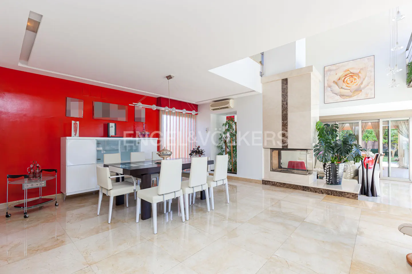 Modern dining room with a dark wood table, white chairs, a bold red accent wall, and a marble fireplace with a large plant.