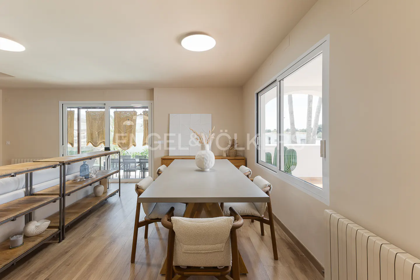 Bright dining room with a long gray table, six chairs, and a white vase. Shelves and a window with a view of cacti are visible.