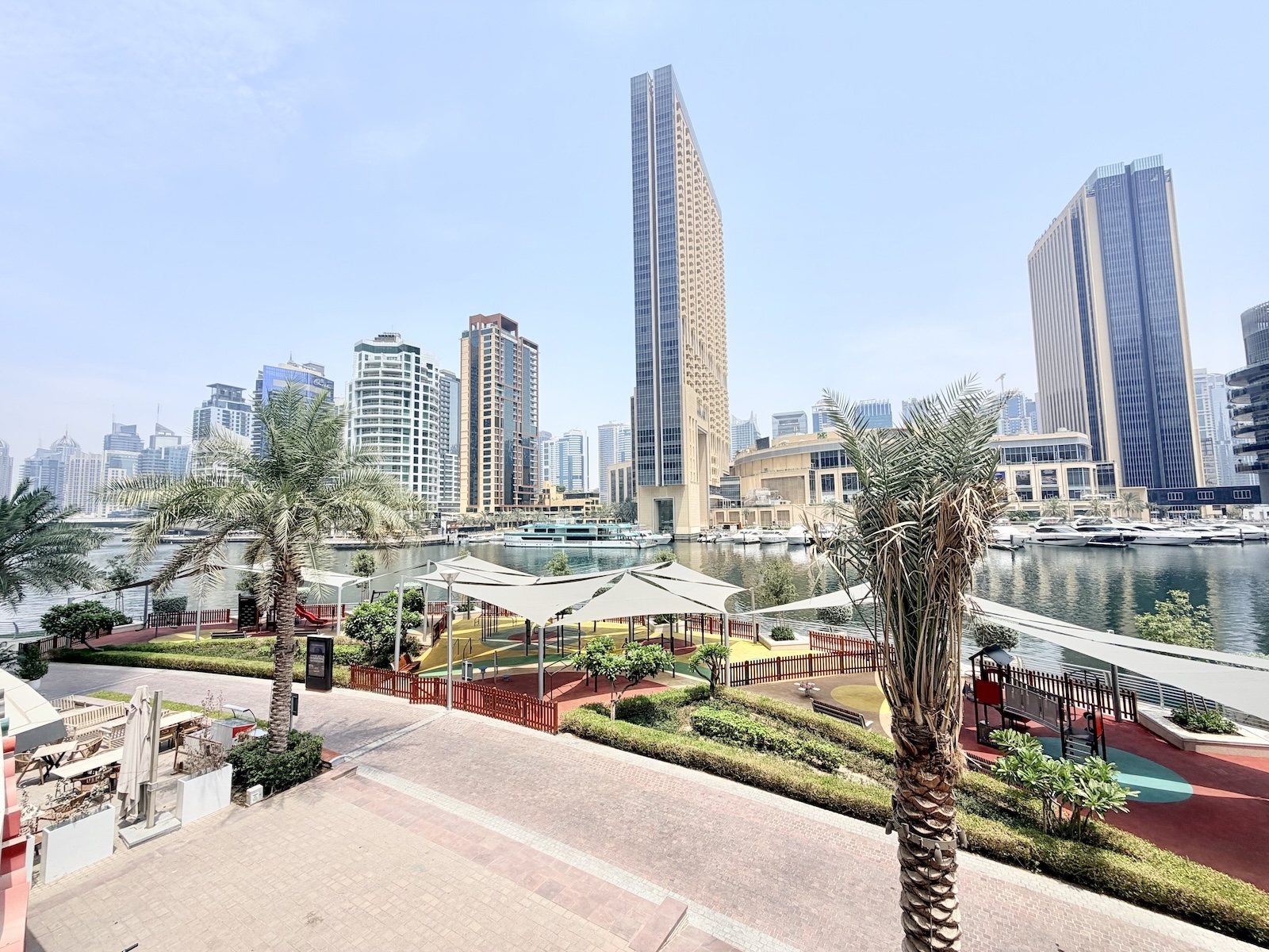 A sunny waterfront park with palm trees, a shaded playground, and a view of modern skyscrapers across the canal.
