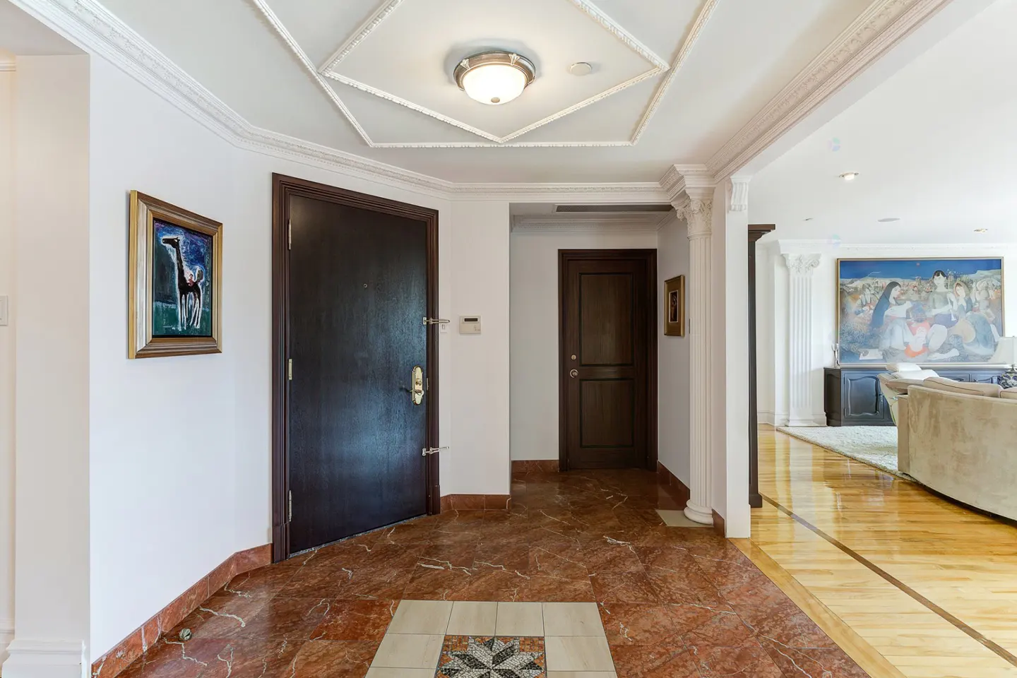 Elegant foyer with red marble floors, dark wood doors, crown molding, and a view into a living room with large wall art.