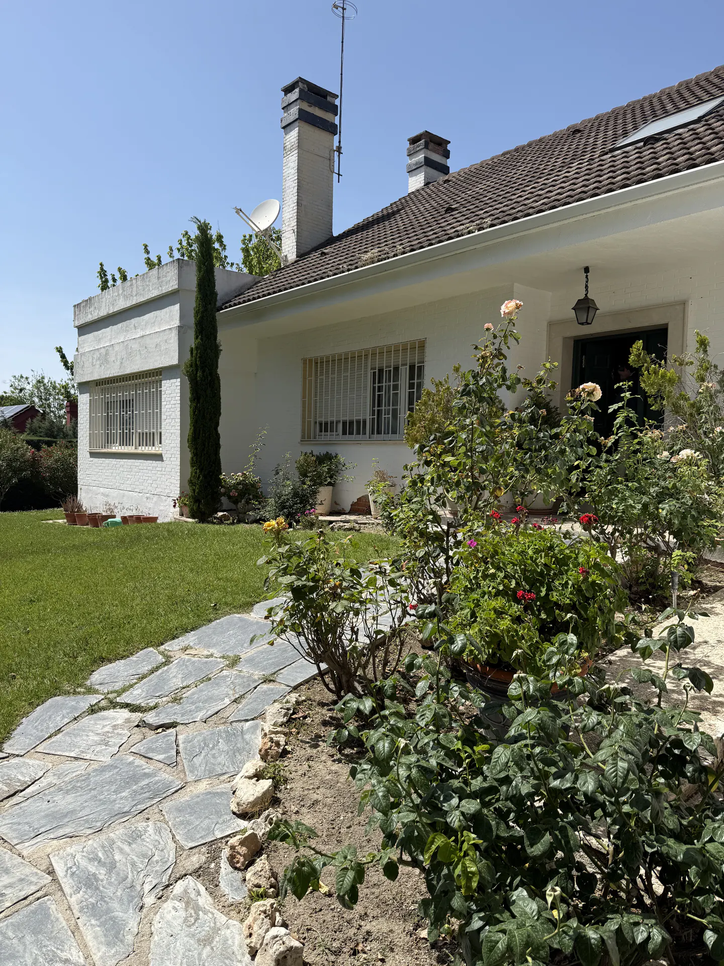 Exterior view of a white single-story house with a stone walkway, green lawn, and flower garden. Chimneys and a satellite dish are visible on the roof.