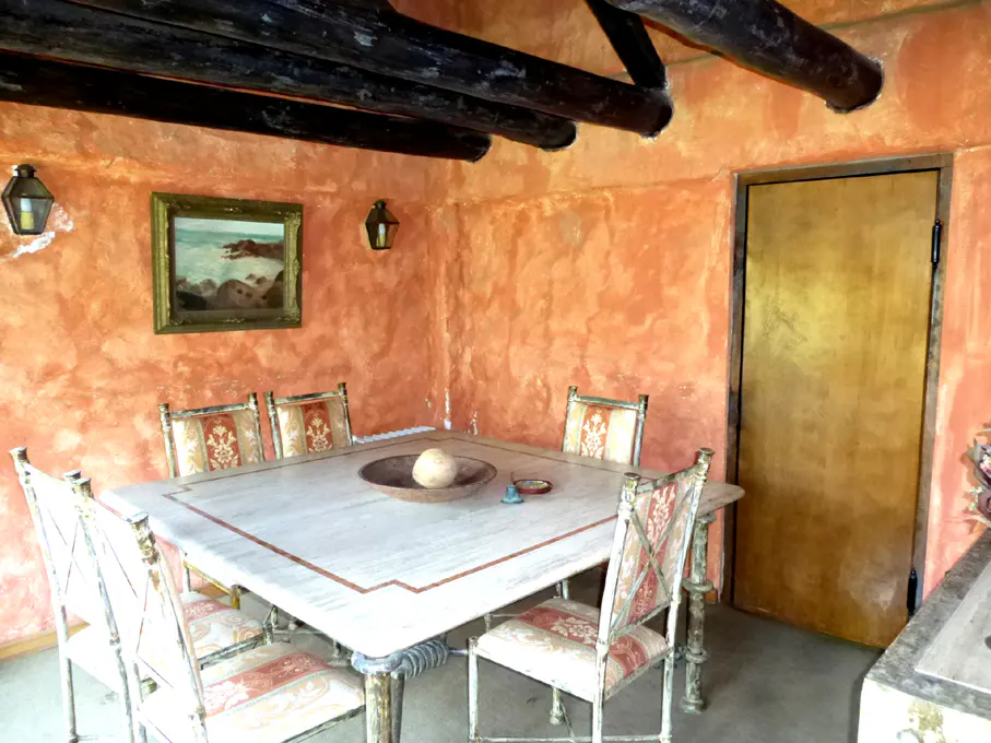 Dining room with orange textured walls, dark wood beams, and a square marble table with six chairs. A landscape painting hangs on the wall.