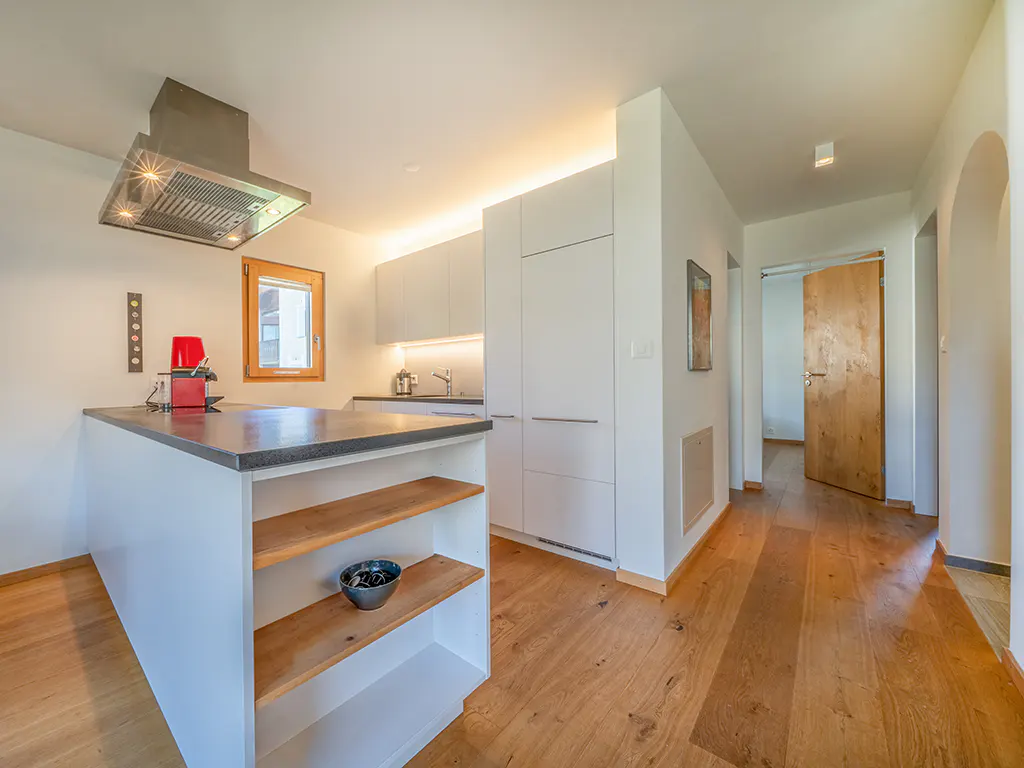 Modern kitchen with white cabinets, a central island with wooden shelves, and light wood flooring leading to a hallway.