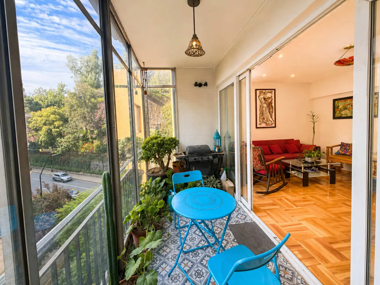 Enclosed balcony with blue table and chairs, grill, and plants. Living room with red sofa visible through sliding glass doors.