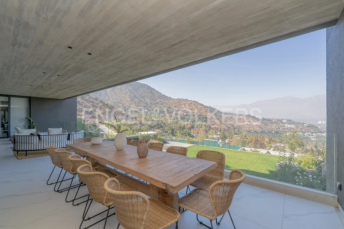 Outdoor patio with a long wooden table, wicker chairs, and mountain views. Modern design with a concrete ceiling and glass railing.