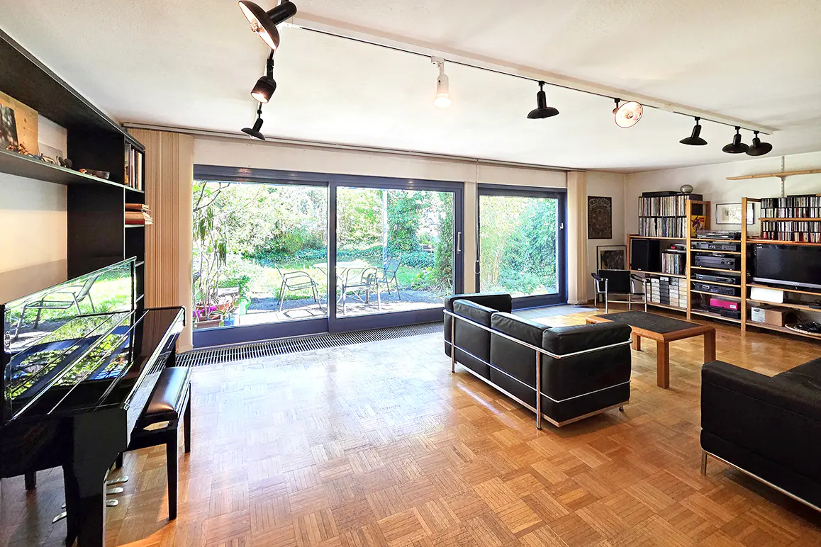 Spacious living room with parquet floors, black leather sofas, a grand piano, and large sliding doors opening to a garden.