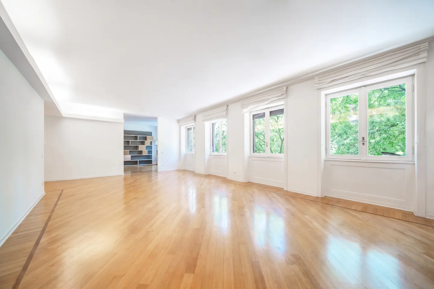 Bright, empty room with polished light wood floors, white walls, and large windows overlooking green trees.