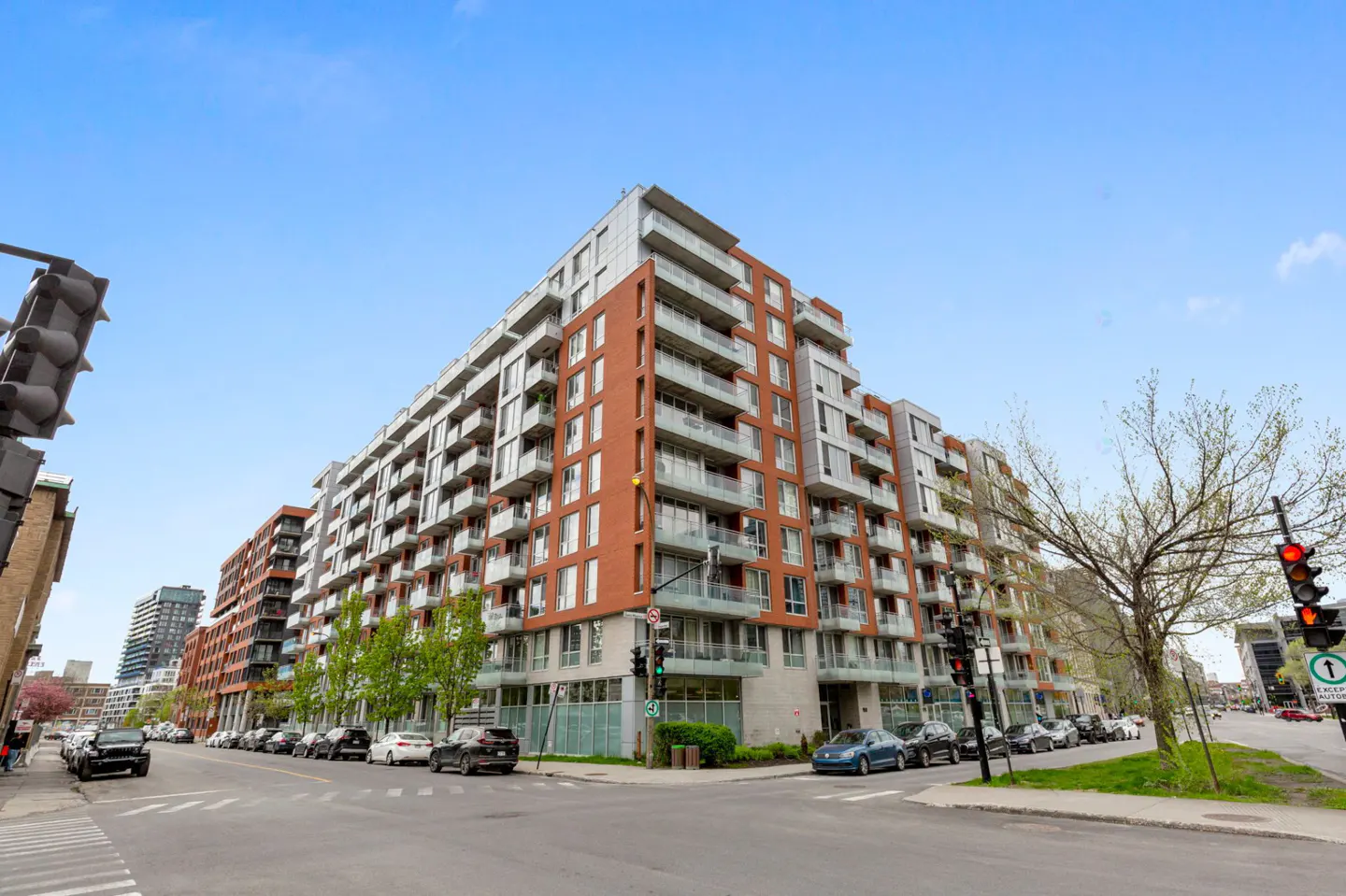 A modern mid-rise apartment building with red brick and grey panels on a city street corner under a clear blue sky.