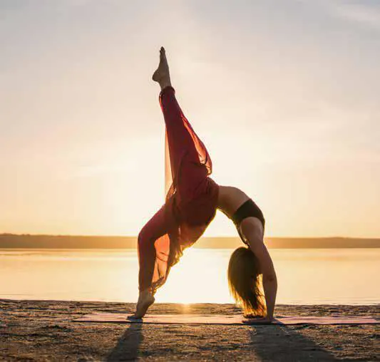 A woman in a black top and red pants does yoga on a mat at sunset by the water.