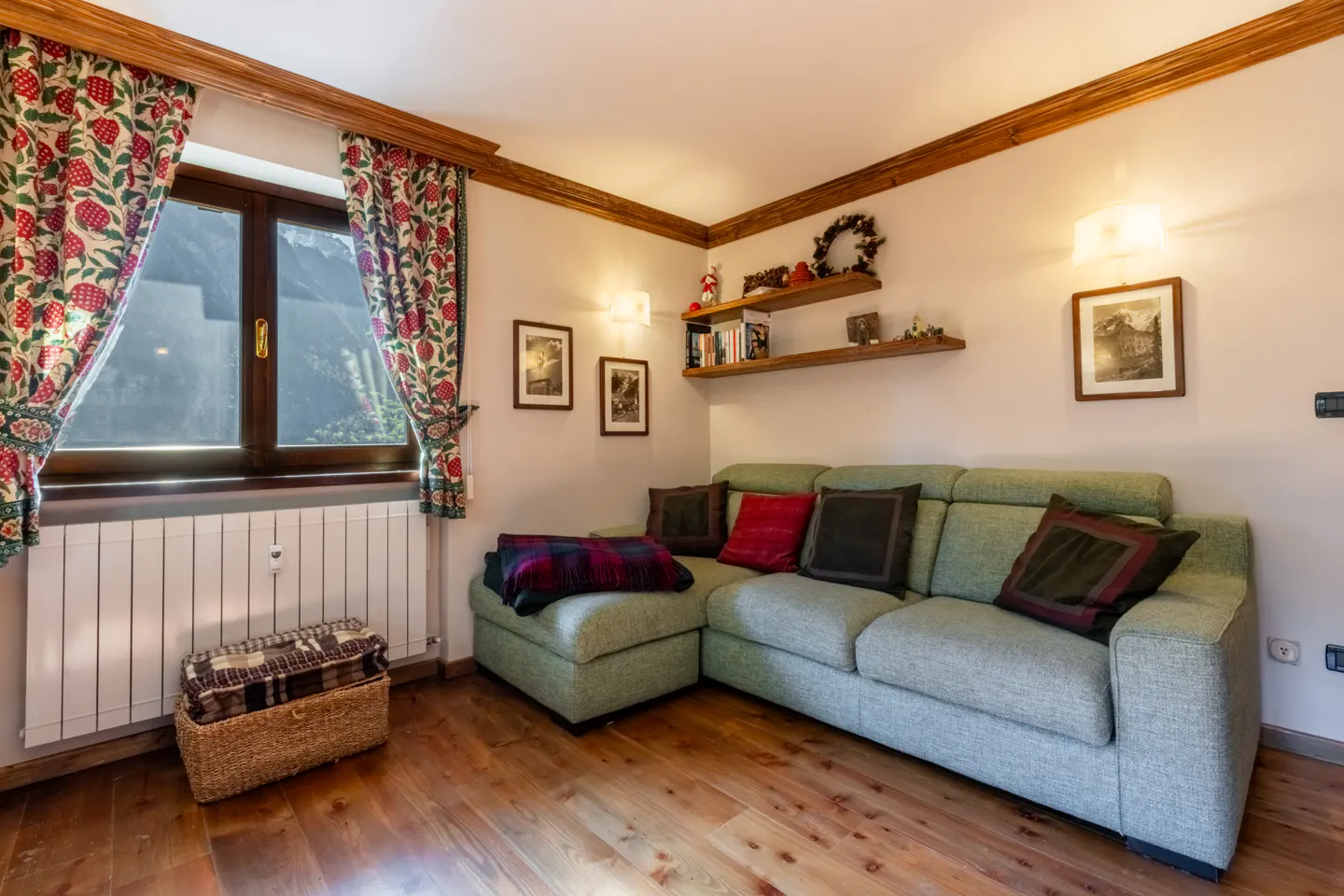 Living room with a light green sectional sofa, wooden floors, and a window with floral curtains. Shelves with books and decor are on the wall.