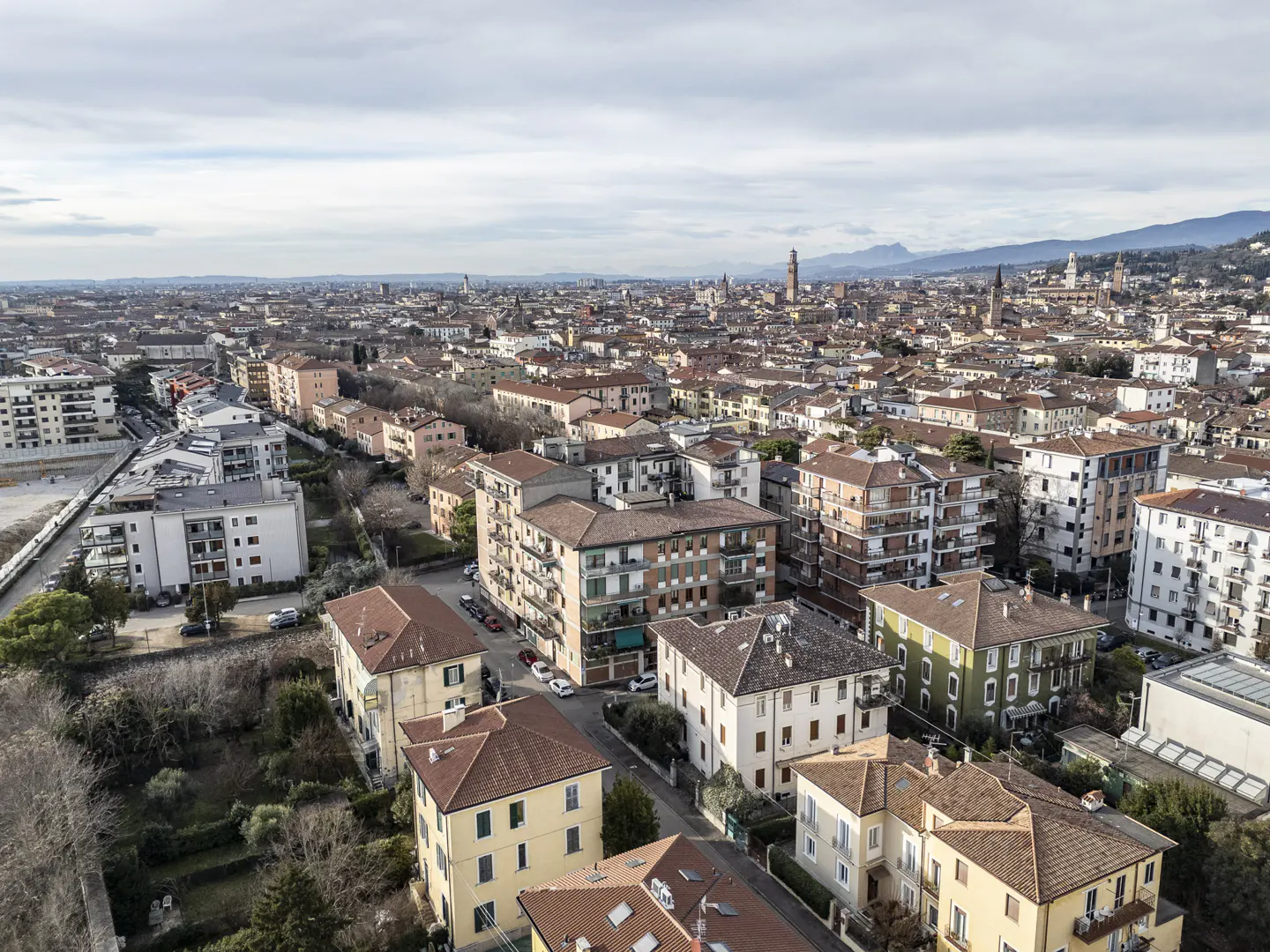 Aerial view of Verona, Italy, showing buildings with red tile roofs and distant mountains under a cloudy sky.