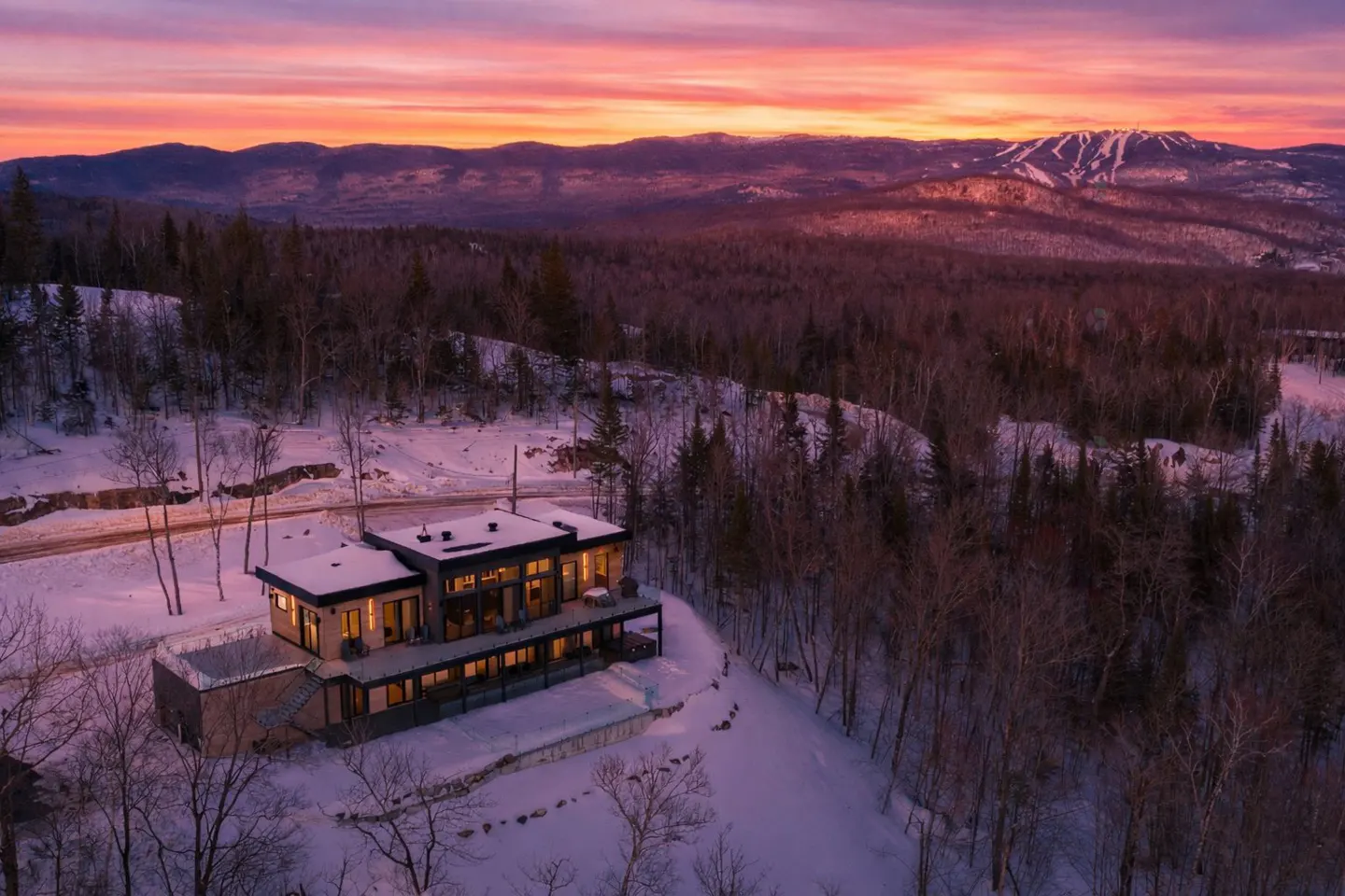 Modern home in snowy landscape at sunset. Two-story house with large windows, surrounded by trees and mountains under a colorful sky.