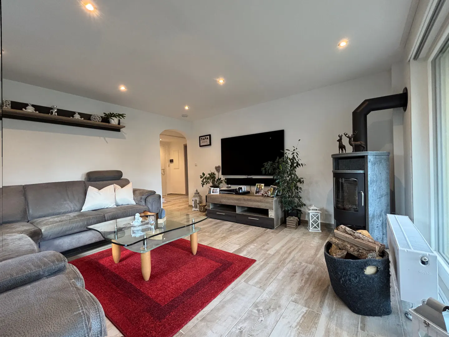 Modern living room with a grey sectional sofa, glass coffee table on a red rug, flat-screen TV, and a wood-burning stove.