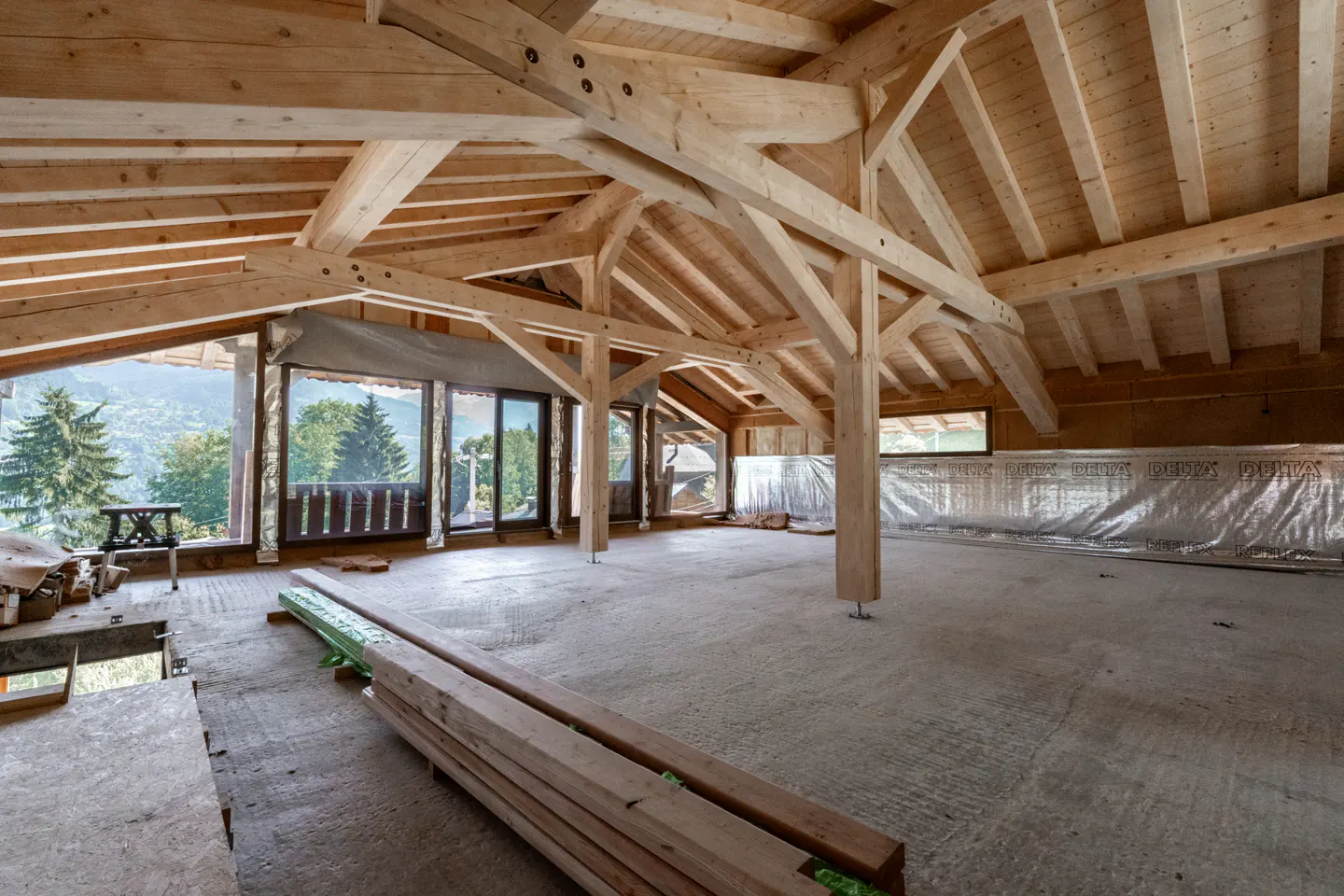 Unfinished attic with exposed wooden beams and large windows overlooking a green, mountainous landscape.