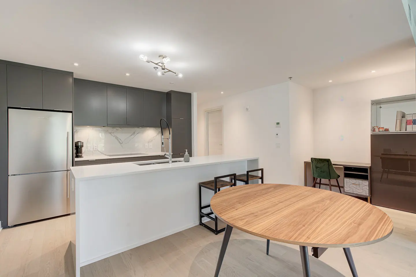 Bright, modern kitchen with gray cabinets, stainless steel fridge, white countertops, and a round wooden table in the foreground.