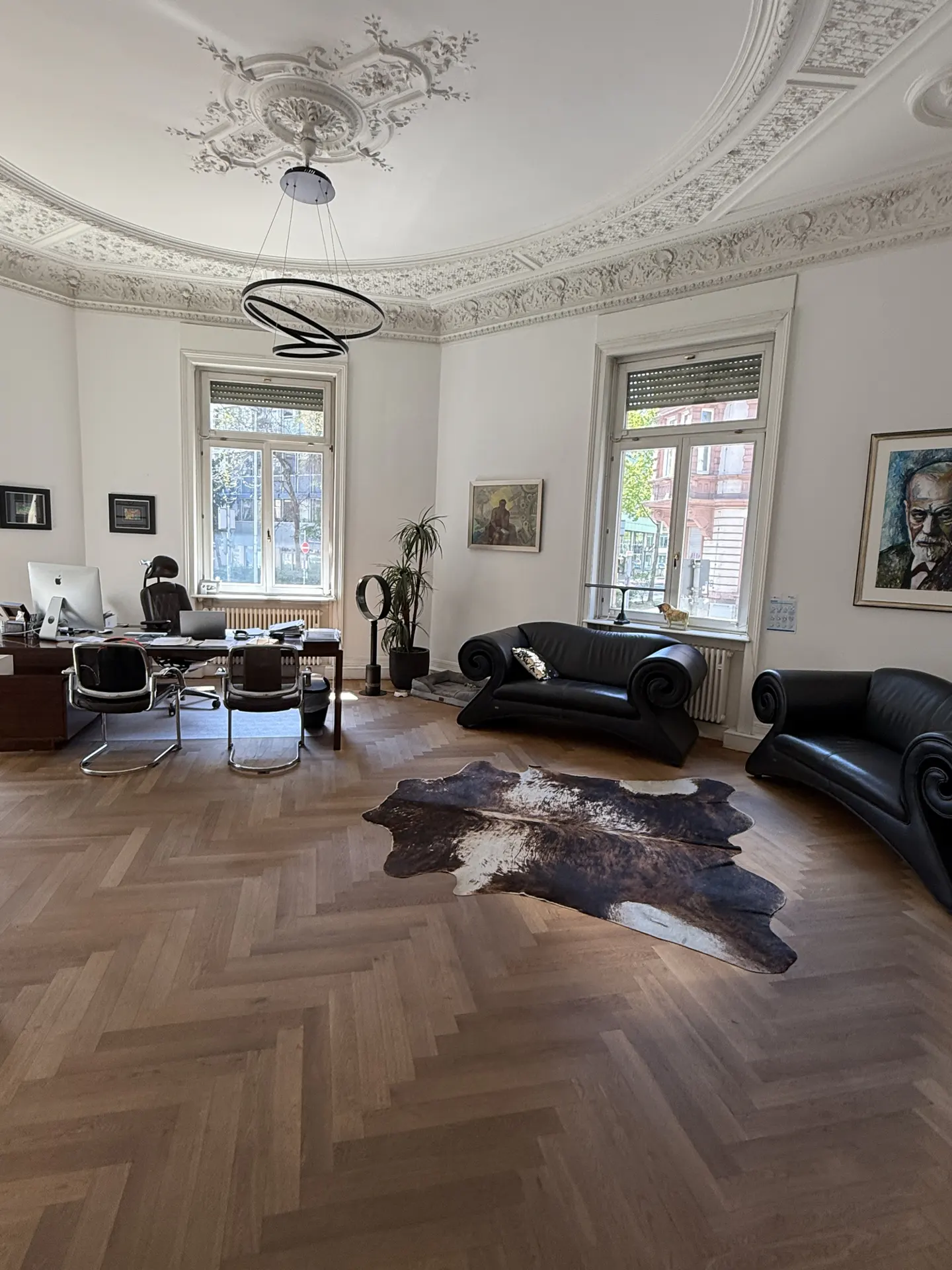 Bright office with herringbone wood floors, black leather sofas, and a cowhide rug. A desk and chairs sit near a window.
