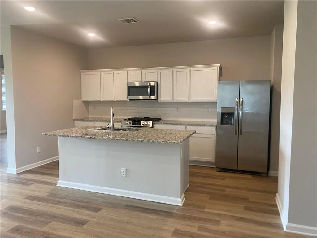 A bright kitchen with white cabinets, stainless steel appliances, and a granite island. Wood-look flooring adds warmth.