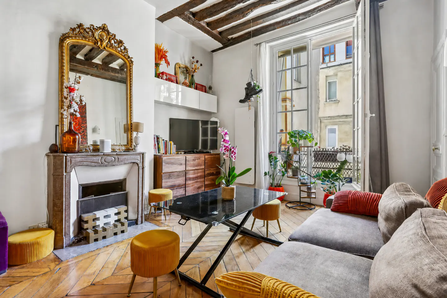 Bright living room with herringbone wood floors, fireplace with gold mirror, grey sofa, and open window to a balcony with plants.