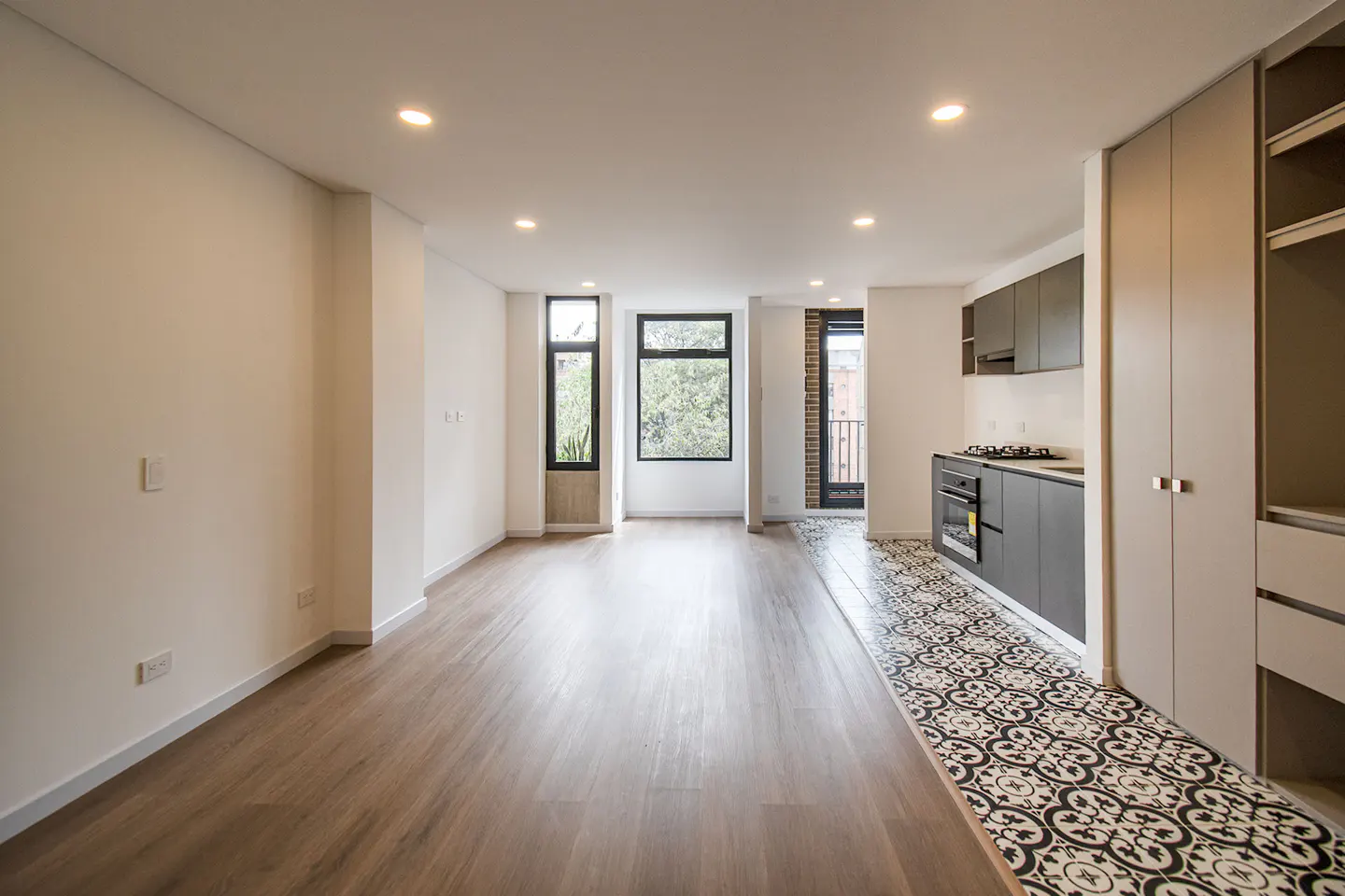 Bright, modern apartment with wood floors, white walls, and black-framed windows. A gray kitchen with patterned tile is visible.
