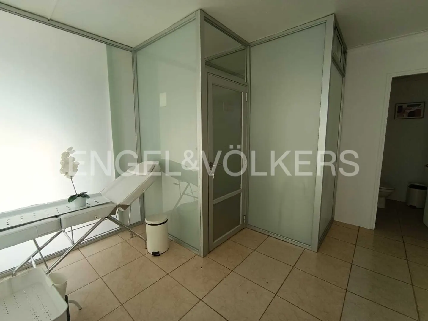A bright room with a white massage table, white orchid, and frosted glass partitions. A bathroom is visible in the background.