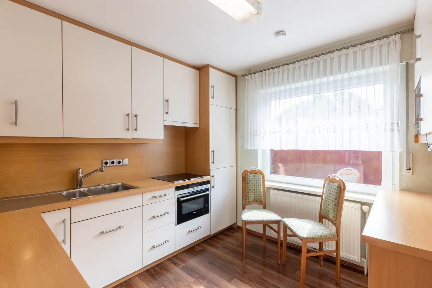 Bright kitchen with white cabinets, wood countertops, and dark wood floors. Two chairs sit by a window with sheer white curtains.