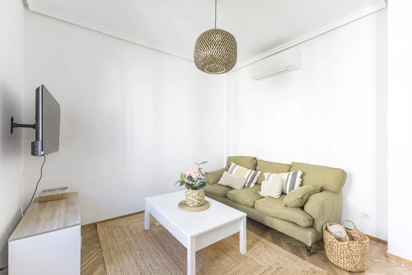 Living room with a green sofa, white walls, a white table with flowers, and a woven rug. A TV hangs on the wall.