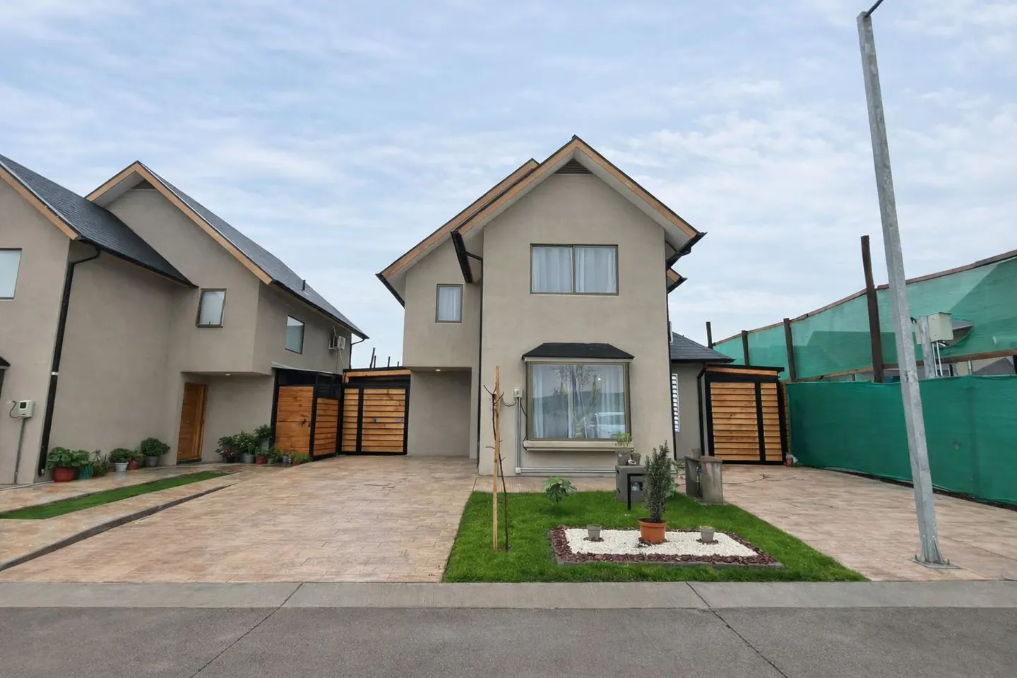 Two-story beige house with a dark roof and wood accents on a cloudy day. A small garden is in front.