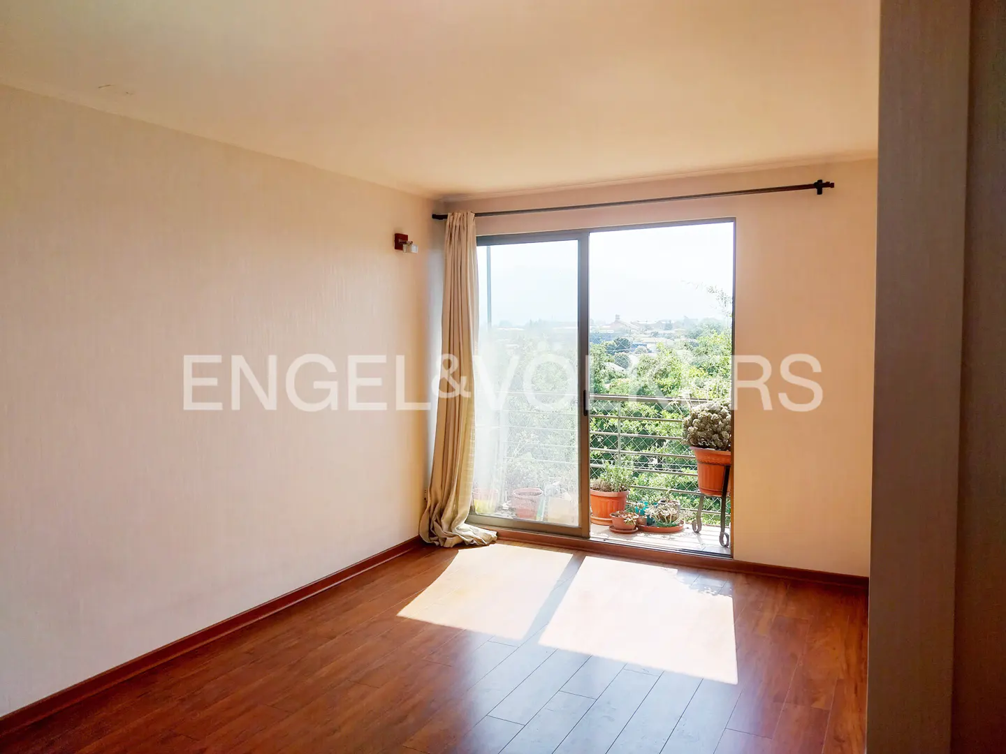 A bright, empty room with wood floors and a sliding glass door to a balcony with plants.