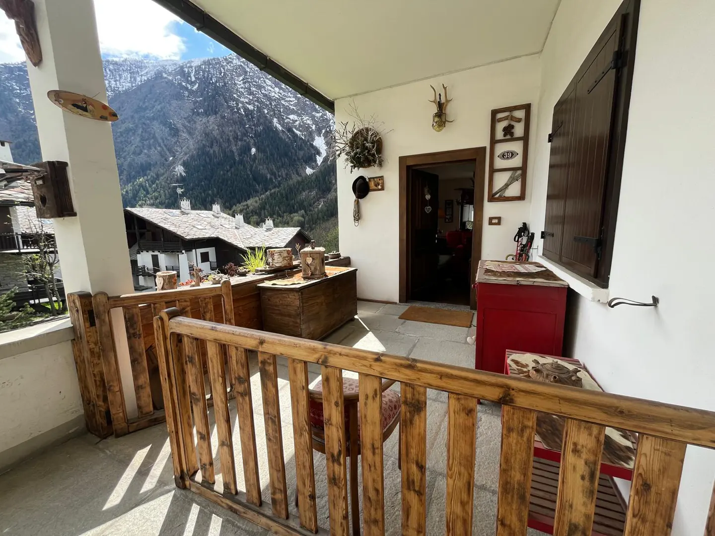 Covered porch with wooden railing, chests, and mountain view. A red cabinet and decorations adorn the white walls.