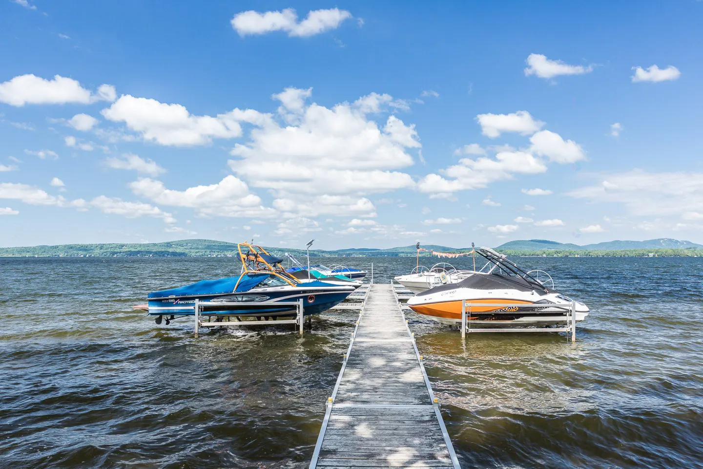 A wooden dock leads to boats on lifts in a lake under a blue, cloudy sky.