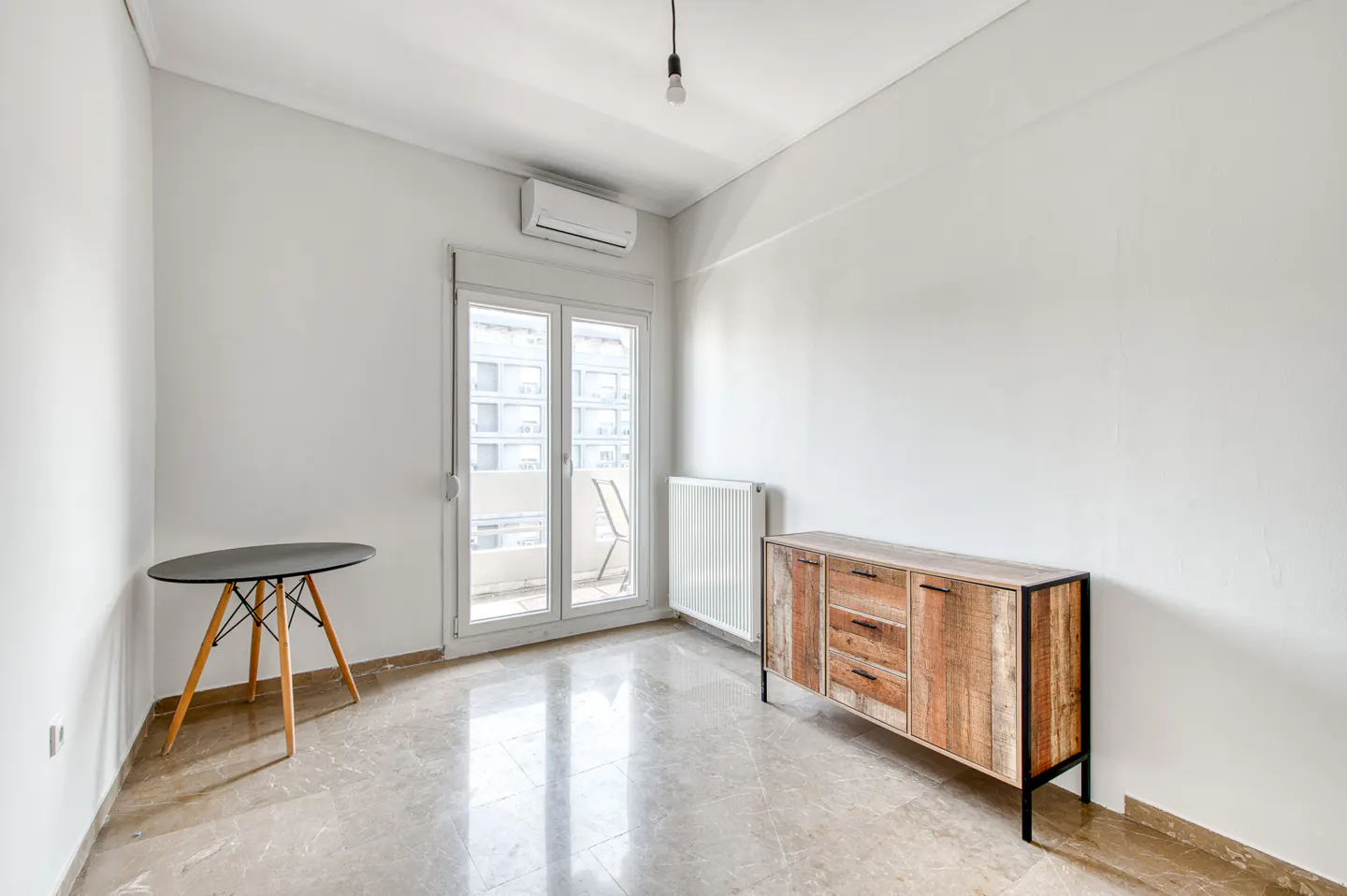 Bright, empty room with marble floors, white walls, and a balcony. A wooden cabinet and small table furnish the space.