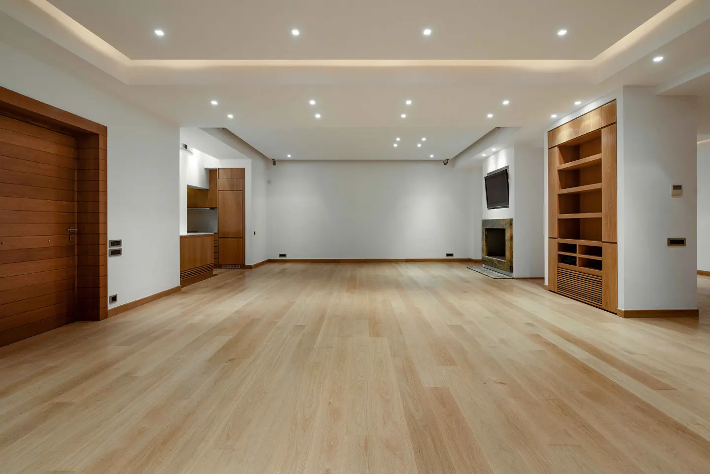 Bright, empty living room with light wood floors, white walls, and recessed lighting. A fireplace and built-in shelves are on the right.