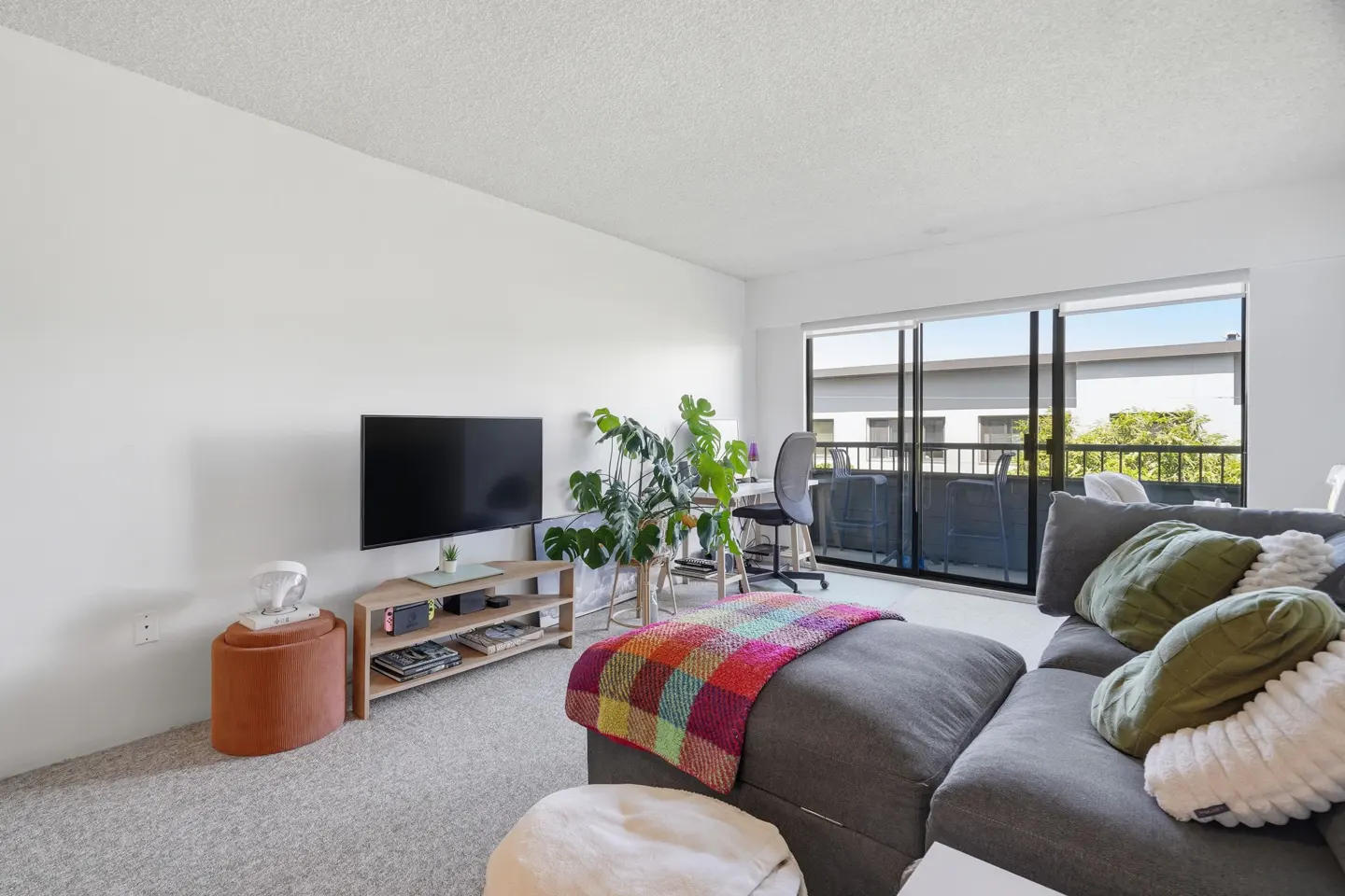 Bright living room with gray sectional sofa, colorful blanket, TV, and balcony access. White walls and carpet.