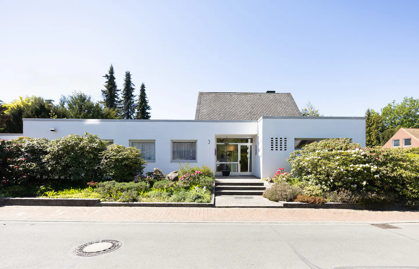 A modern, one-story white house with a gray roof, surrounded by lush green bushes and colorful flowers. A gray street is in the foreground.