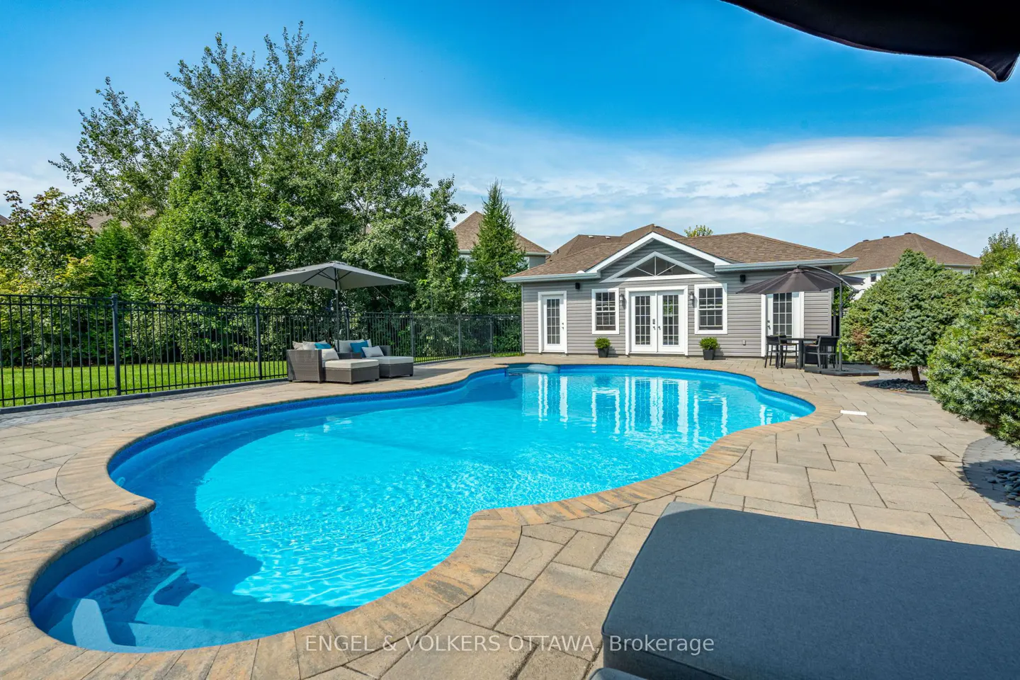 Outdoor pool with blue water, stone patio, gray pool house with white doors, and lounge furniture under a gray umbrella.