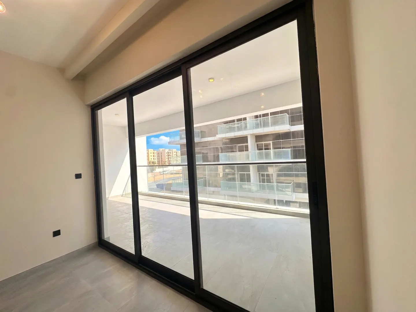 A bright room with a large black-framed sliding glass door leading to a balcony with a view of a modern building.