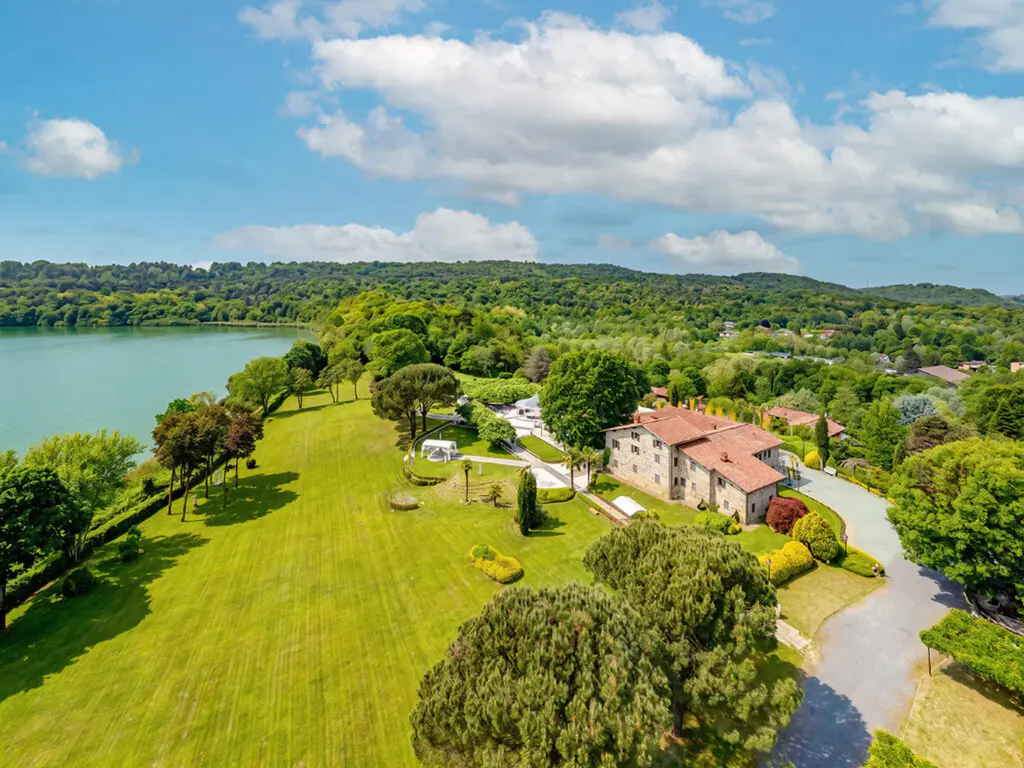 Aerial view of a stone villa with a red roof, surrounded by lush green lawns, trees, and a lake under a blue sky.