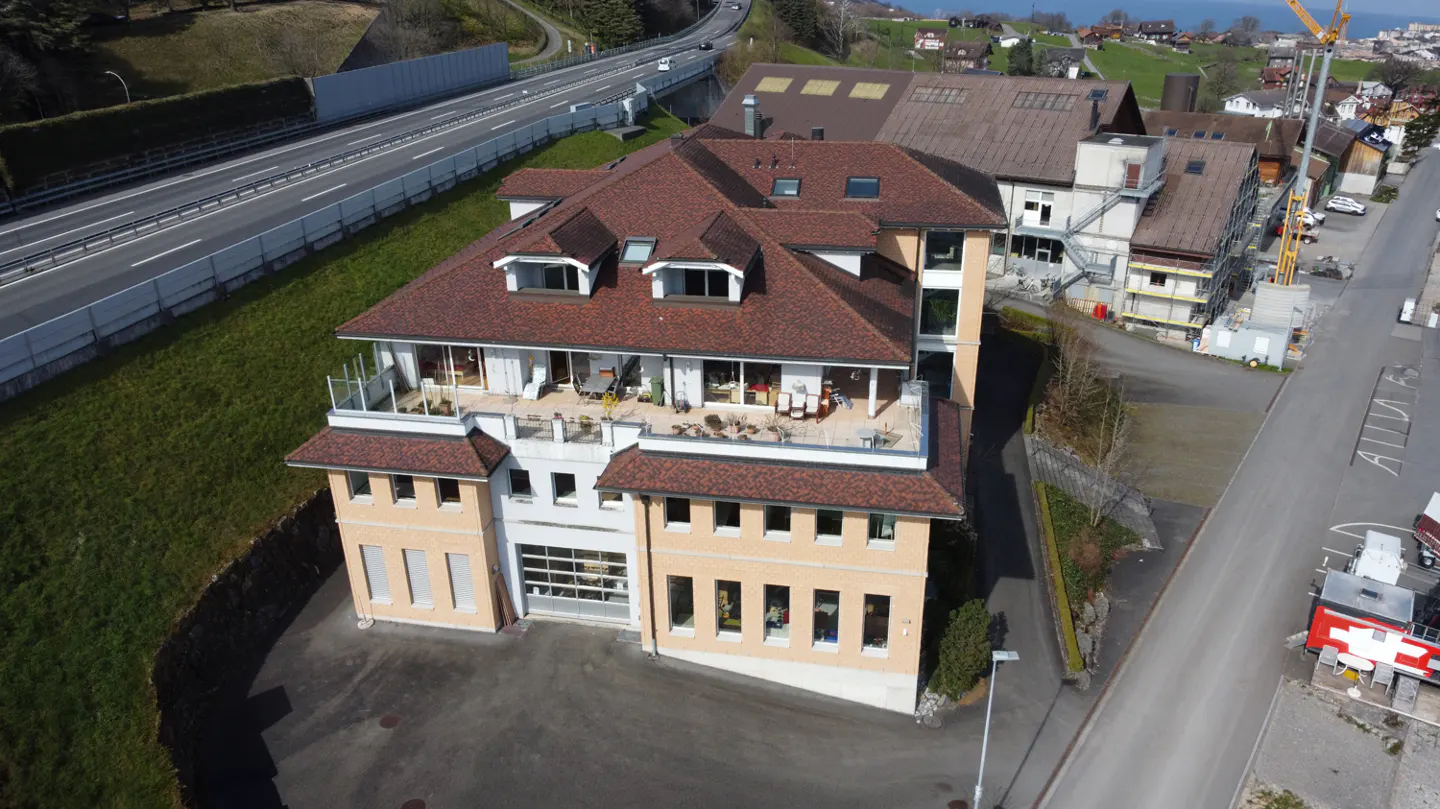 Aerial view of a multi-story building with a brown roof and a beige facade, featuring a rooftop terrace and a garage door. A highway runs behind the building.