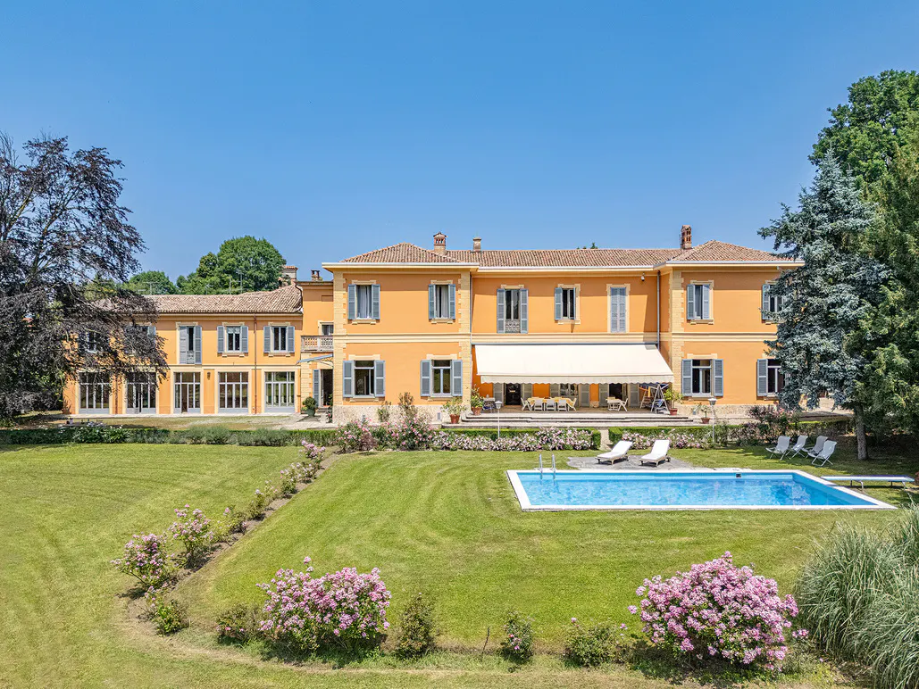 Exterior view of a large, orange two-story house with a pool, lawn, and trees under a clear blue sky.