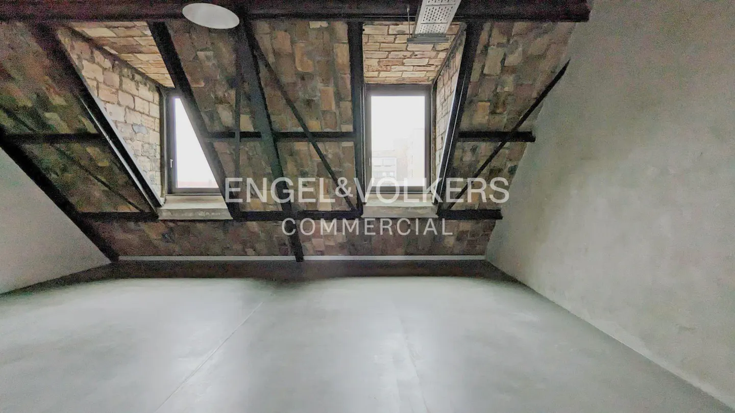 Attic room with brick walls, black beams, and two windows. The floor is gray concrete. "Engel & Volkers Commercial" is superimposed on the image.
