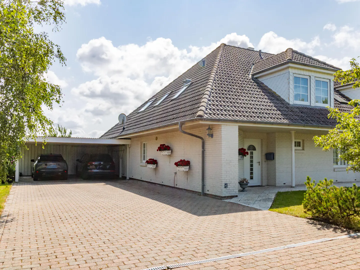 Exterior view of a white brick house with a gray tiled roof, a brick driveway, and two cars parked in a carport.