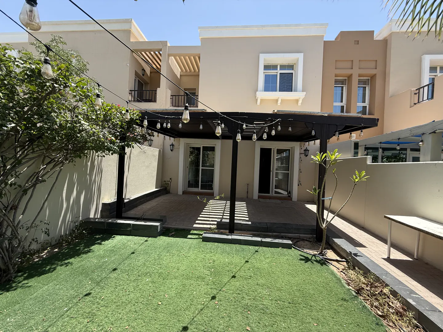 Backyard of a beige two-story house with a black pergola, string lights, and green artificial grass.