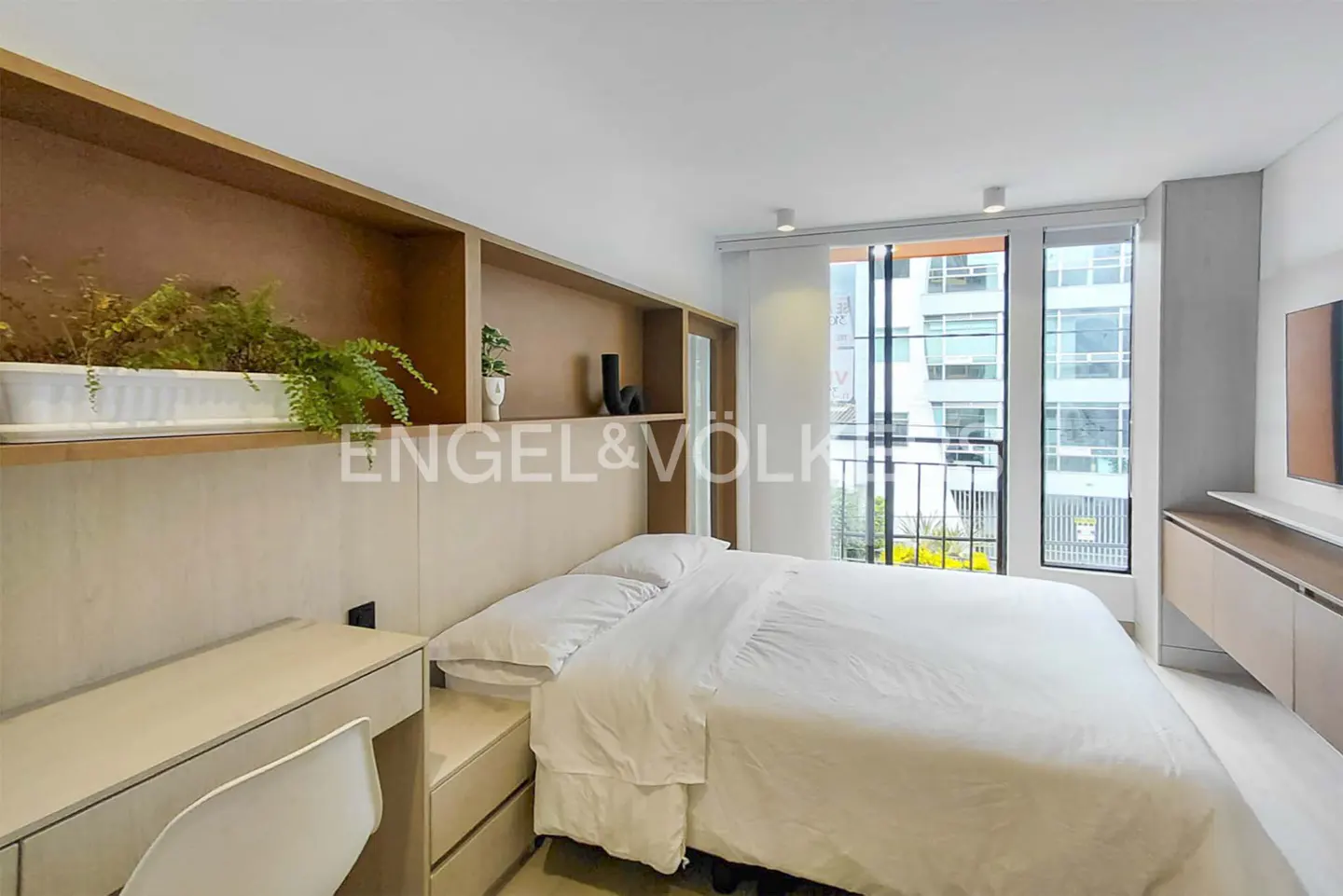 Modern bedroom with a white bed, light wood desk, and built-in shelving featuring a green plant, overlooking a city street.