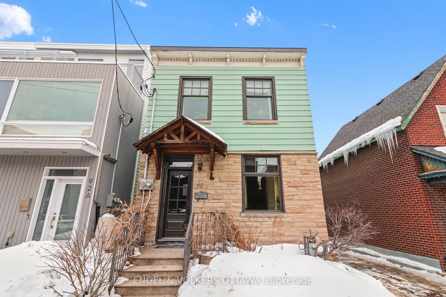 Exterior view of a two-story house with light green siding and a stone facade, set against a blue sky with snow on the ground.