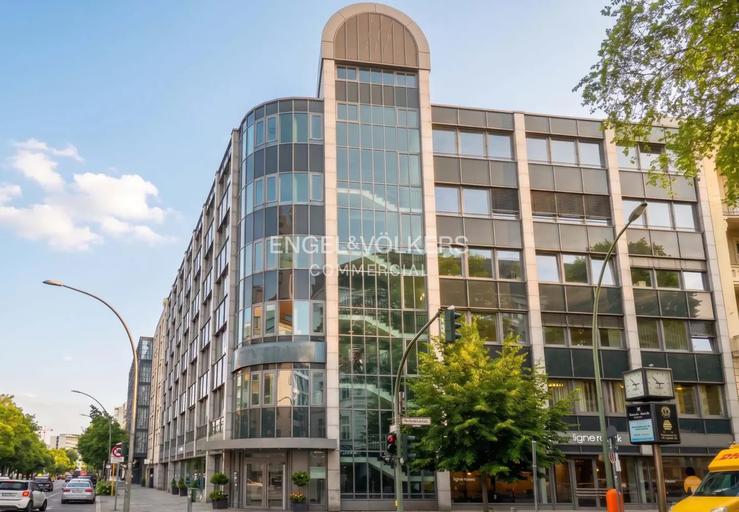 Exterior view of a modern, multi-story commercial building with glass and gray facade, street-side view with trees and street lamps.