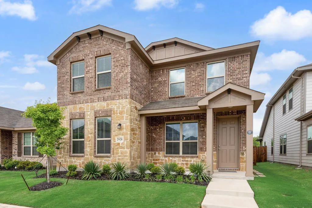 Two-story house with brick and stone facade, brown door, green lawn, and blue sky.