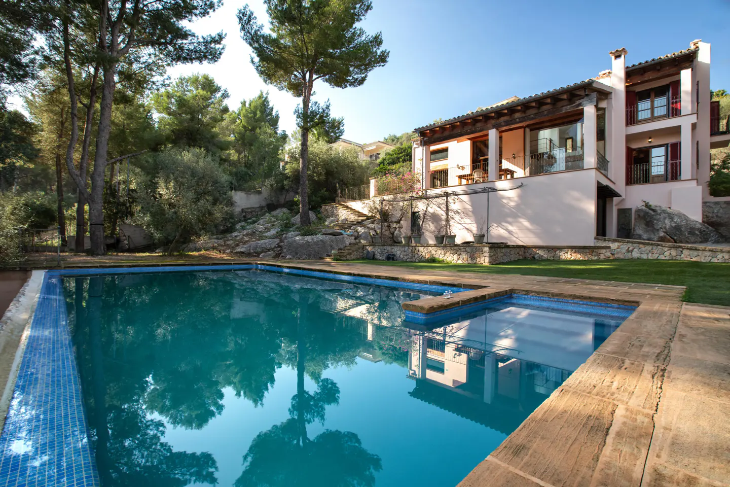 A turquoise pool reflects trees and a two-story pink house with red shutters on a sunny day.