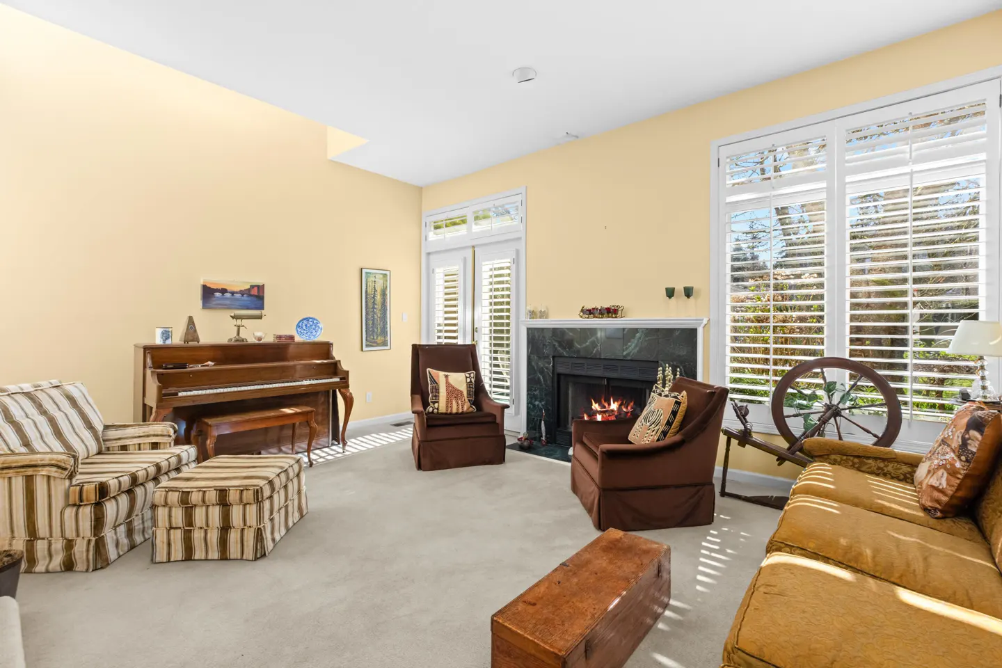 Living room with yellow walls, fireplace, piano, brown chairs, and striped sofa. Sunlight streams through shuttered windows.