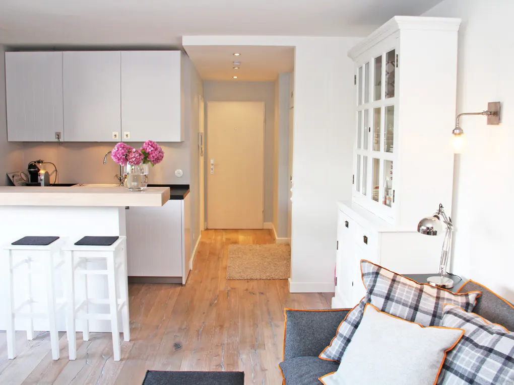 Bright, modern apartment interior with white cabinets, breakfast bar, and stools. A hallway leads to a white door. A gray couch with pillows is in the foreground.