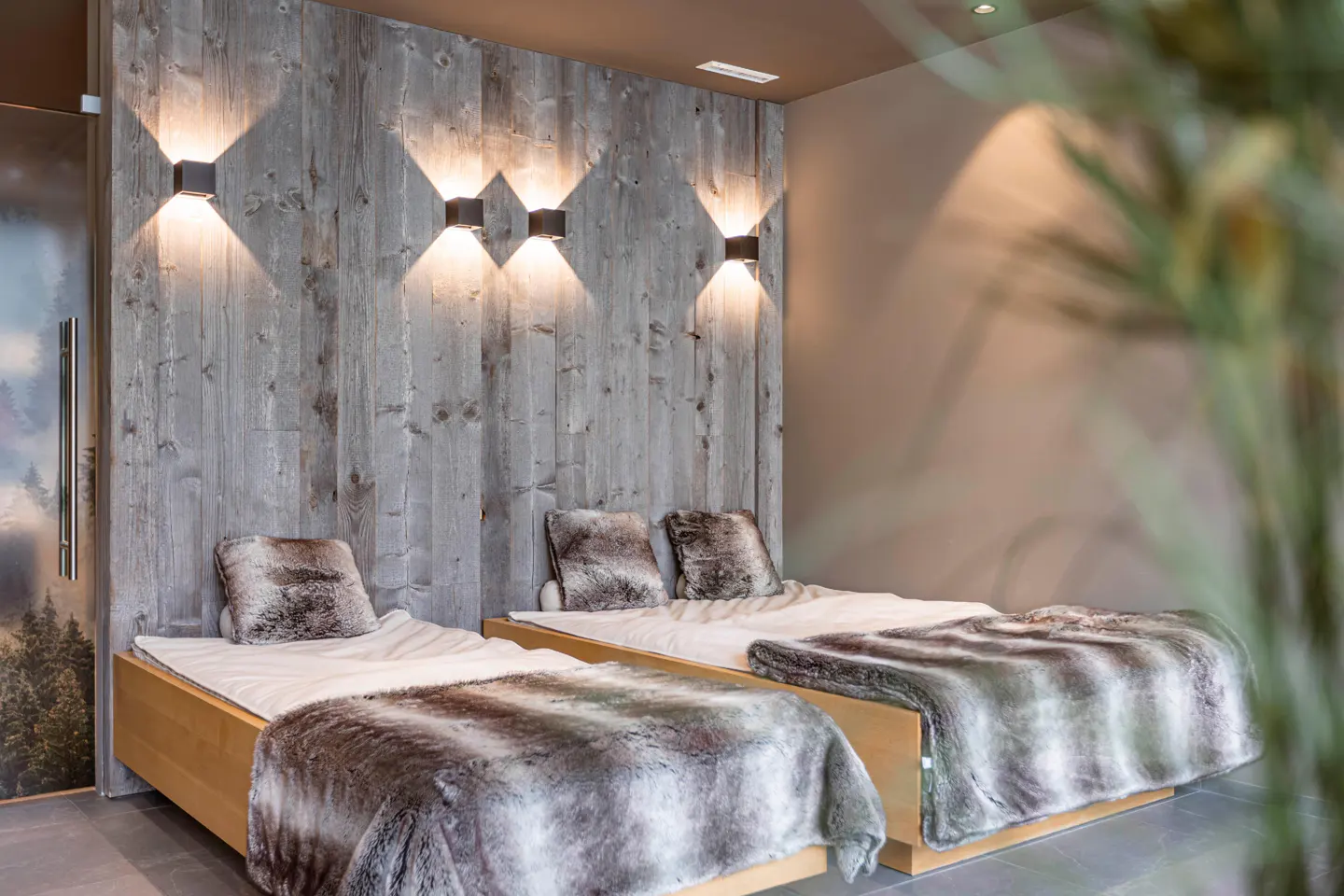 Two twin beds with faux fur blankets against a rustic grey wood-paneled wall with warm, modern wall sconce lighting.